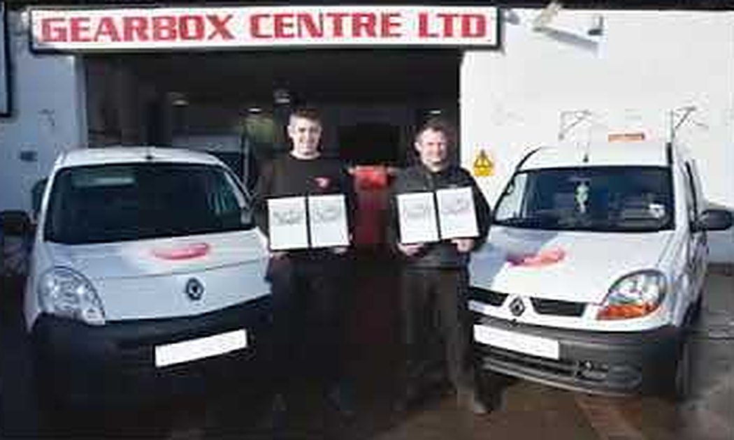 Two Gearbox Centre team members holding framed certificates outside the workshop, under the Gearbox Centre Ltd sign, between two company vans